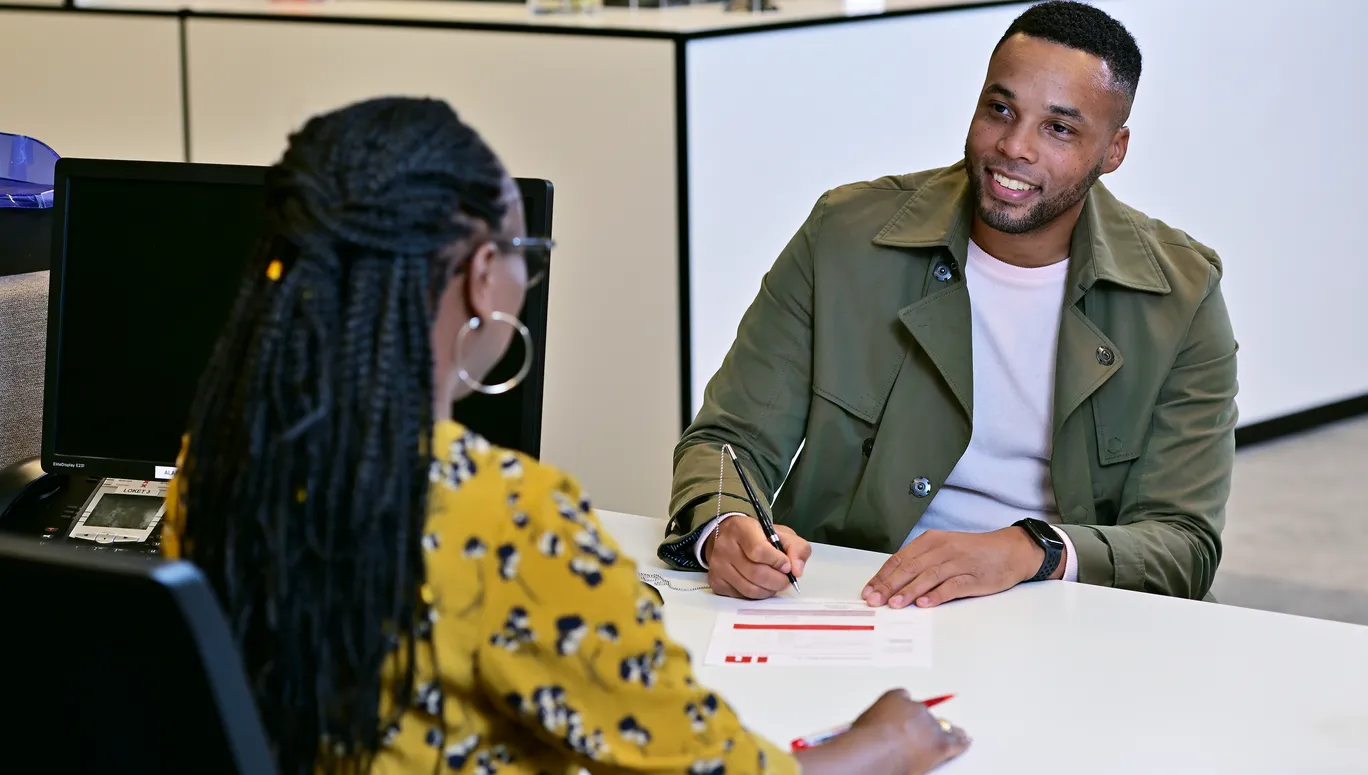 Man sitting at the desk in the INAmsterdam offices, seen from the front, talking to employee, seen from the back